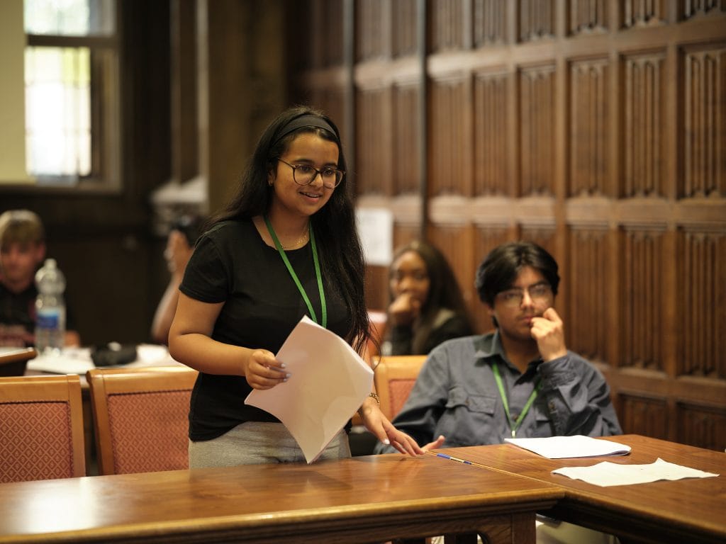 A girl stands up and speaks, holding papers in one hand. A boy sits next to her.